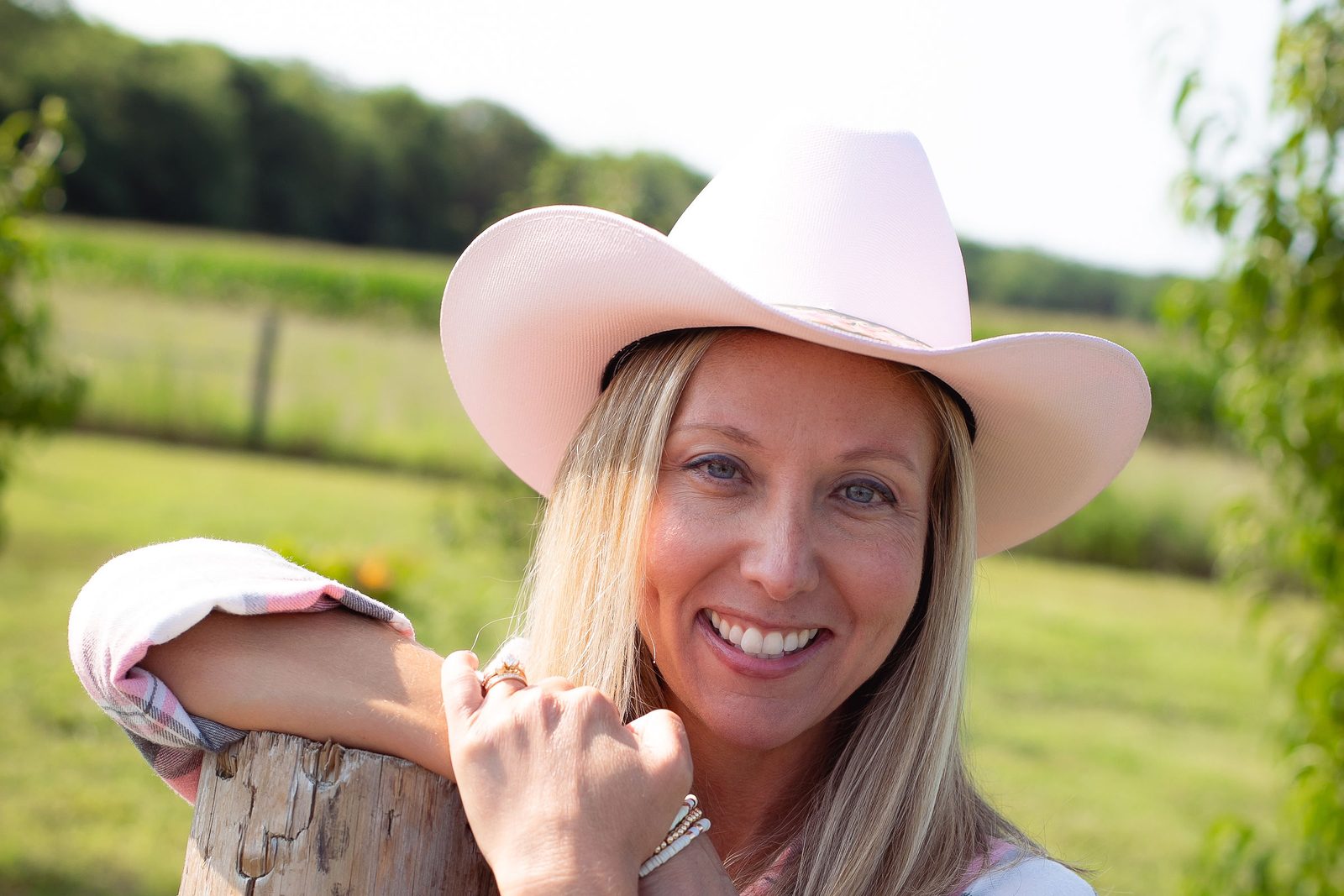 Crystal Joy in a pink cowboy hat, smiling and leaning on a fence post in a sunlit country field